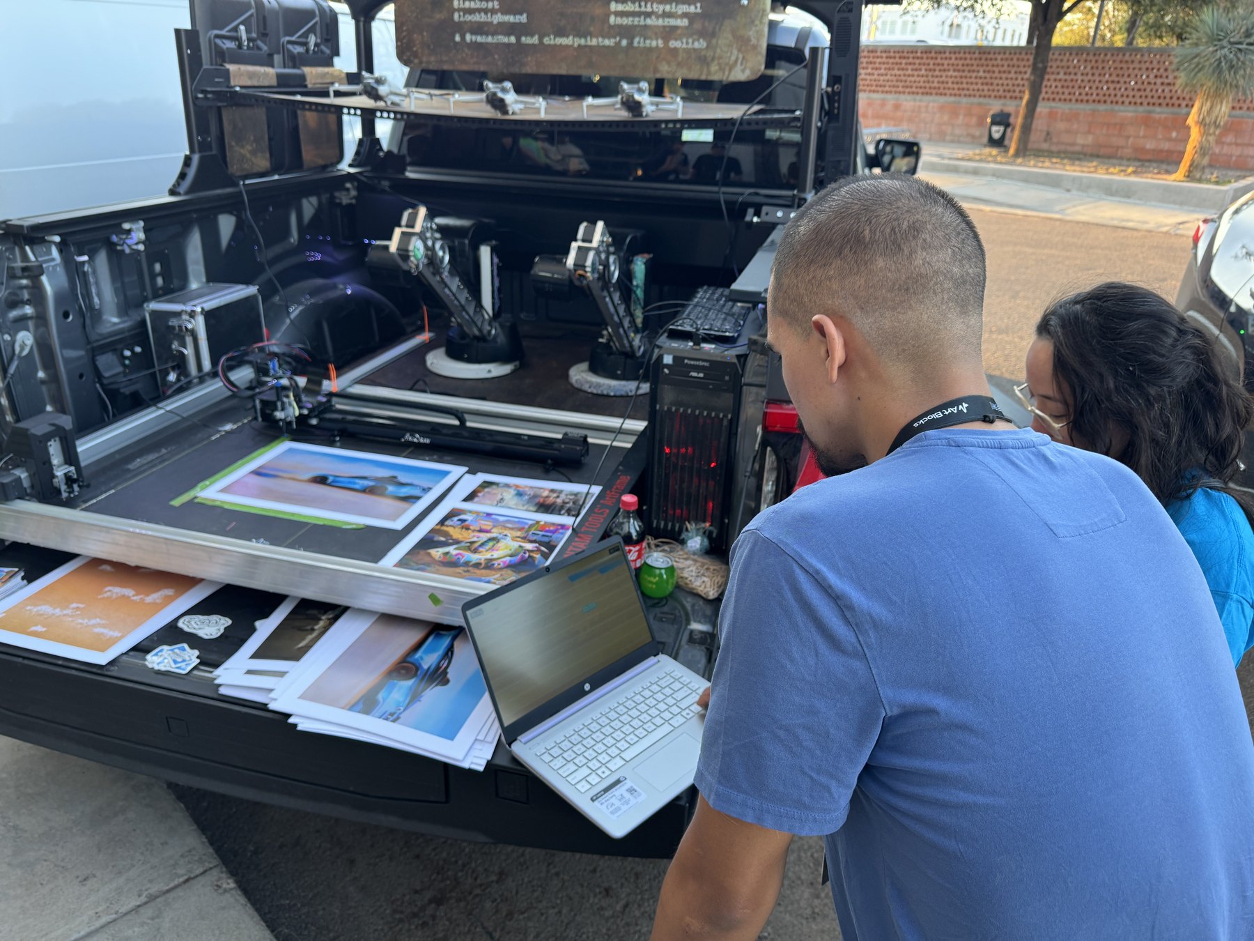 The open tailgate of the cloudpainter truck: a pen plotter drawing on a print, two robot arms, a tower PC, and prints laid out, with the artist billing board above